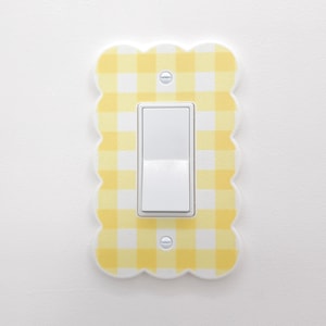 May include: A decorative light switch cover with a yellow and white gingham pattern. The cover has a scalloped edge and a white toggle switch in the center. The cover is mounted on a white wall.