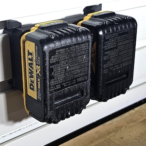 Może przedstawiać: Dwa czarne i żółte akumulatory DeWalt 20V Max XR Lithium Ion zamontowane na białej ścianie za pomocą czarnego uchwytu ściennego.