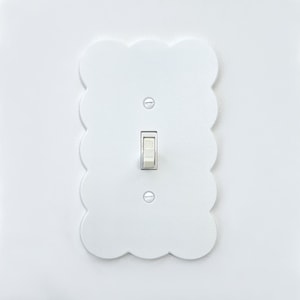 以下が含まれることがあります： 波状の縁取りのある白いライトスイッチカバー。スイッチはオフの位置です。