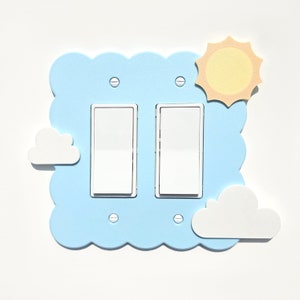 Könnte beinhalten: Eine hellblaue Doppel-Lichtschalterplatte mit einem Wolken- und Sonnenmotiv. Die Platte hat einen gewellten Rand und ist aus dickem Material gefertigt.