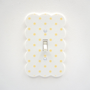 Puede incluir: Una cubierta de interruptor de luz blanca con un diseño de borde festoneado y lunares amarillos. El interruptor de la luz está en el centro de la cubierta, con un interruptor de palanca blanco. La cubierta está montada en una pared blanca.