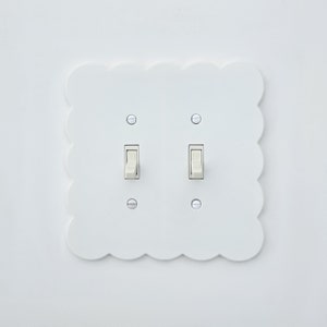 以下が含まれることがあります： 白い壁に、2つの白いロッカー付きのトグルスイッチがついた白いライトスイッチプレートが取り付けられています。プレートは波状の縁取りがされており、白い壁に取り付けられています。