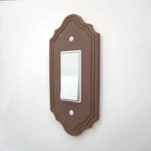 May include: A brown decorative light switch cover with a white toggle switch. The cover has a scalloped edge design and is mounted on a white wall. The switch is in the on position.
