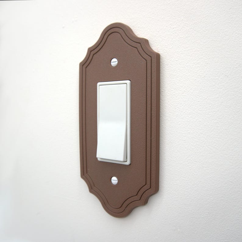 May include: A brown decorative light switch cover with a white toggle switch. The cover has a scalloped edge design and is mounted on a white wall. The switch is in the on position.