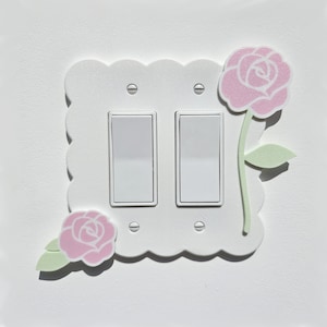 Può includere: Copertura per interruttore della luce bianca con bordo a festoni e due interruttori a levetta. La copertura è decorata con fiori di carta rosa e verdi.