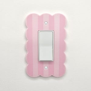 Könnte beinhalten: Eine hellrosa und weiß gestreifte Lichtschalterabdeckung mit gewelltem Rand. Die Abdeckung hat einen weißen Kippschalter in der Mitte und ist an einer weißen Wand montiert. Zwei Schrauben befestigen die Abdeckung.