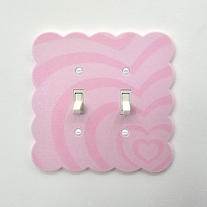 May include: A pink light switch cover with a wavy, heart-shaped design. The cover has a scalloped edge and two white light switches. The design features shades of pink and white, creating a whimsical aesthetic.