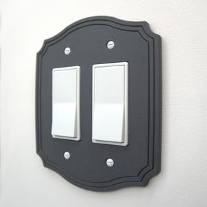 May include: A decorative, dark gray double light switch cover with a scalloped edge. The cover has two white light switches and is secured with four silver screws. The design is simple and elegant, suitable for various interior styles.