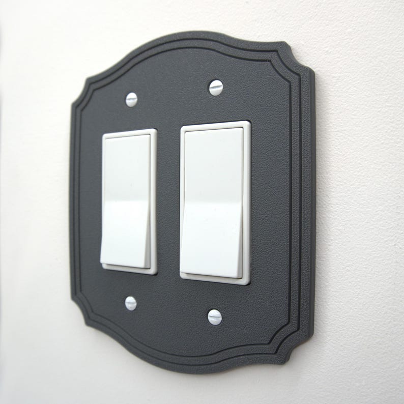 May include: A decorative, dark gray double light switch cover with a scalloped edge. The cover has two white light switches and is secured with four silver screws. The design is simple and elegant, suitable for various interior styles.