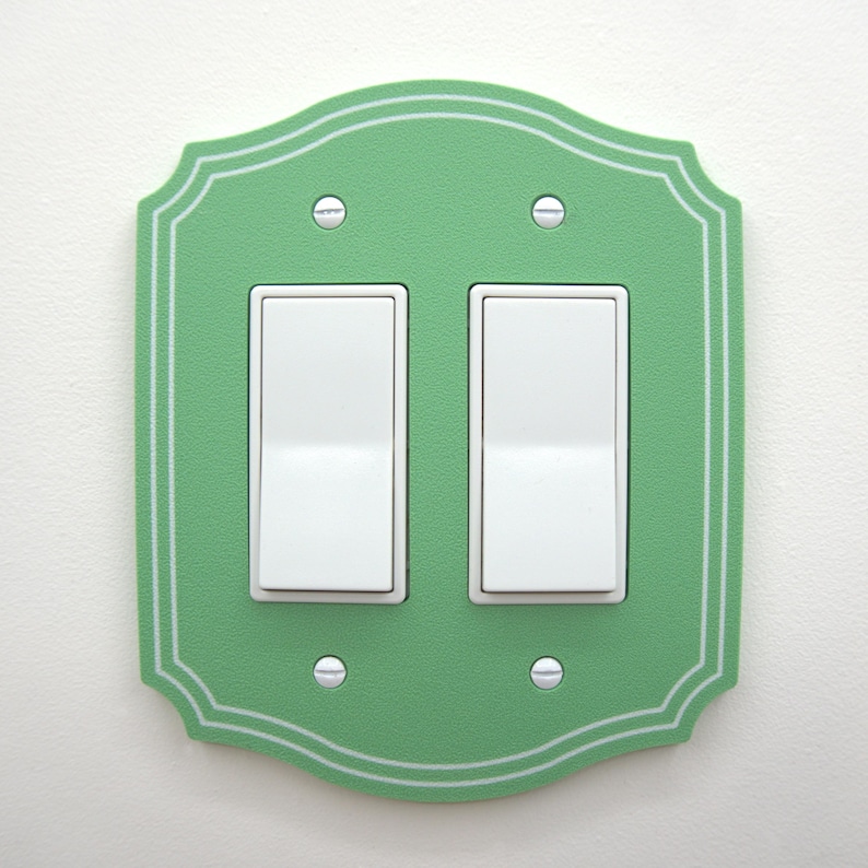 May include: A double rocker light switch wall plate with a scalloped edge and a matte green finish. The plate features a thin white double-line border detail and is secured by four small silver-colored screws. Two white rocker switches are centered.
