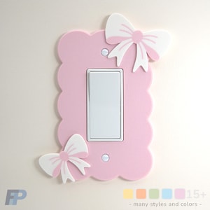 Op de afbeelding: Een roze lichtschakelaar cover met een geschulpte rand en twee witte strikken met roze centra. De cover heeft een witte schakelaar en is gemonteerd op een lichtgekleurde muur. Tekst onderaan luidt "15+ - many styles and colors -."