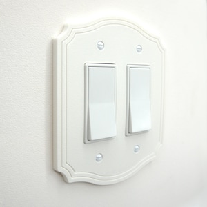 May include: White double light switch plate with a decorative, scalloped edge. The plate has two rectangular light switches and is mounted on a white wall. The screws are visible and match the color of the plate.