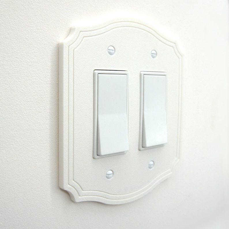 May include: White double light switch plate with a decorative, scalloped edge. The plate has two rectangular light switches and is mounted on a white wall. The screws are visible and match the color of the plate.