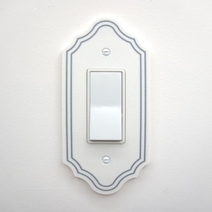 May include: A white light switch with a decorative, scalloped edge plate. The plate has a gray border and is mounted on a white wall. The switch is in the on position.