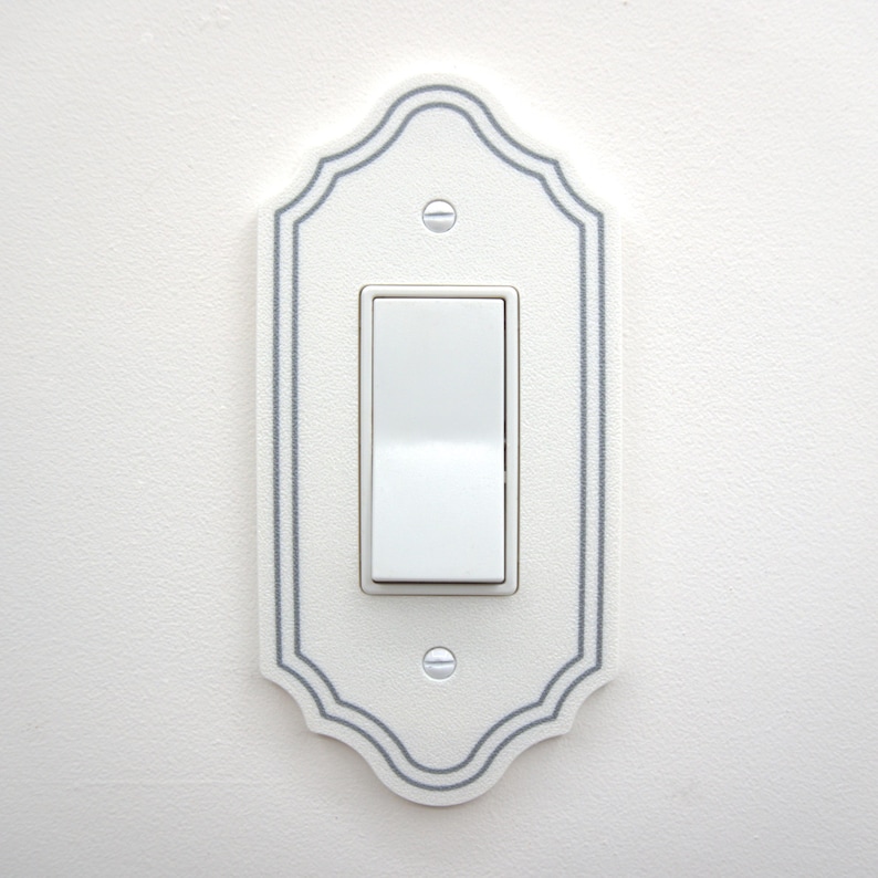 May include: A white light switch wall plate with a decorative scalloped edge and a thin gray border line. The rectangular plate features a central white rocker switch and is secured to the wall with two visible white slotted screws.