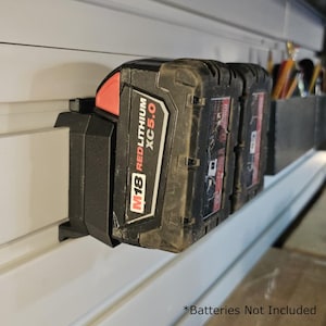 May include: Black M18 Red Lithium XC5.0 battery pack mounted on a wall. The battery pack is black with red accents and white text. The text "M18 Red Lithium XC5.0" is visible. The image also shows a storage container for tools.