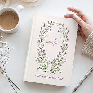 Może przedstawiać: Biały notatnik w twardej oprawie z akwarelową ilustracją fioletowego wieńca kwiatowego otaczającego imię Amelia, z napisem Letters to my daughter wydrukowanym na fioletowo u dołu, w towarzystwie kubka kawy i srebrnego długopisu.