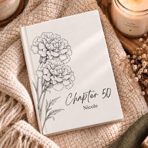 Op de afbeelding: Een wit hardcover boek met een zwarte lijntekening van twee anjers aan de linkerkant. De tekst "Chapter 50" is in een handschriftlettertype gedrukt boven de naam "Nicole" in een eenvoudig schreeflettertype op de voorkant van de kaft.