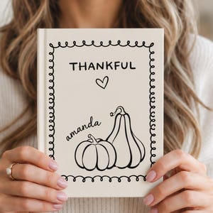 May include: A cream-colored hardcover journal featuring a black line-art border of loops. The cover displays the word THANKFUL, a small heart, the name amanda in script, and two black-outlined pumpkins of different shapes centered on the front.