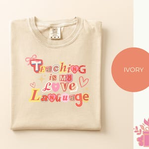 May include: Ivory-colored t-shirt with the text "Teaching is my Love Language" in a colorful, playful font. The design includes hearts, stars, and a bow. The shirt is folded neatly on a light background.