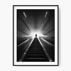 May include: Black and white photograph of a person at the top of a long escalator, ascending into a bright, illuminated tunnel. The image is framed in a black border.