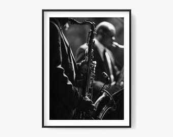May include: Black and white framed photograph of a jazz musician playing a saxophone. The musician is wearing a suit and is in the foreground, with another musician in the background. The image is a close-up shot, focusing on the saxophone and the musician's hands.