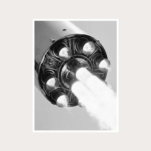 May include: A black and white photograph of a rocket engine base featuring a circular arrangement of multiple thrusters. Two central thrusters are actively emitting bright, dense plumes of exhaust smoke, while the surrounding nozzles remain inactive.
