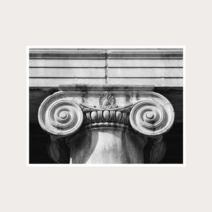 Puede incluir: Una fotografía arquitectónica en blanco y negro de un capitel de columna jónica. El capitel de piedra presenta dos volutas prominentes a los lados, un adorno central de palmeta y una moldura de ovas, sosteniendo un entablamento horizontal.