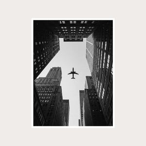 Puede incluir: Fotografía en blanco y negro de un avión a reacción volando entre rascacielos altos. La perspectiva es desde el suelo hacia arriba, enfatizando la altura de los edificios y la silueta del avión contra el cielo. La imagen es un estudio de contrastes.
