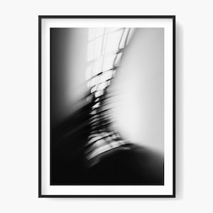 Op de afbeelding: Zwart-wit abstracte kunstafdruk in een zwarte lijst. De afbeelding toont vage lijnen en vormen, met een focus op licht en schaduw. De compositie suggereert architecturale elementen, mogelijk een raam of een gevel.