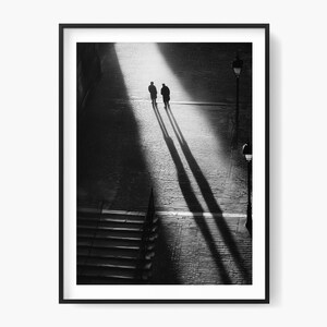 Op de afbeelding: Zwart-wit ingelijste print met twee figuren die over een geplaveide straat lopen, met lange schaduwen achter hen. Een lantaarnpaal en trappen zijn ook zichtbaar. De afbeelding is genomen vanuit een hoog standpunt.