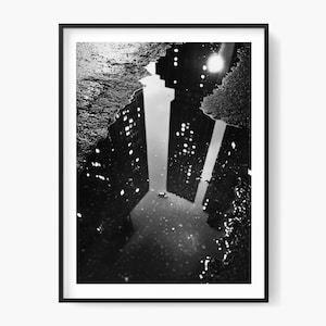 Puede incluir: Fotografía en blanco y negro enmarcada de un reflejo de la ciudad en un charco. La imagen captura los imponentes rascacielos y sus ventanas iluminadas reflejadas en el agua, creando una composición simétrica. El marco es negro.