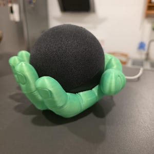 Op de afbeelding: Een zwarte, bolvormige slimme luidspreker rust in een heldergroene, 3D-geprinte handvormige houder. De houder staat op een donkergrijze ondergrond, met een keuken op de achtergrond.