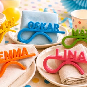 Könnte beinhalten: Ein Set aus fünf bunten Serviettenringen aus Kunststoff, jeder mit einem Namen in erhabenen Blockbuchstaben. Die Namen sind Leon in Gelb, Oskar in Blau, Mats in Grün, Emma in Orange und Lina in Pink. Jeder Ring hat ein geschwungenes Design.