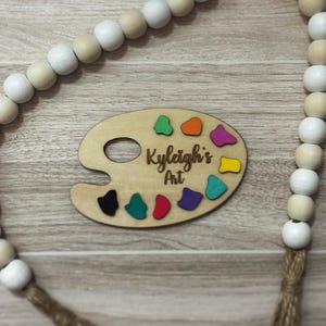 May include: A wooden artist's palette with colorful paint splotches and the text "Kyleigh's Art" is displayed. A beaded garland with white and natural wood beads and a jute tassel is in the background. The palette is a light brown color.