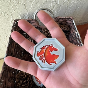 May include: A gray, octagonal pendant featuring a red horse design. The horse is stylized with flowing lines and a curved body. The pendant is held in a hand, with a basket and pinecones in the background.