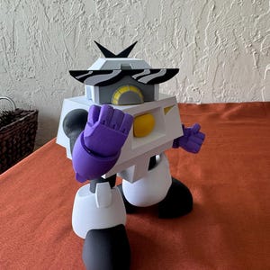 May include: A white, black, and purple robot figurine with a geometric design. The robot has zebra-striped sunglasses, purple hands, and black feet. A yellow sphere is visible in the center of the robot's body.