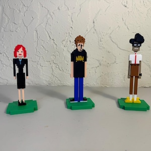 Puede incluir: Tres figuras de arte de píxeles en bases verdes. La figura de la izquierda es una mujer con cabello rojo que lleva un vestido negro y zapatos azules. La figura del medio es un hombre con cabello castaño que lleva una camisa negra con un gráfico de un extraterrestre amarillo, jeans azules y zapatos negros. La figura de la derecha es un hombre con cabello negro que lleva un sombrero negro, una camisa blanca con una raya roja, pantalones marrones y zapatos amarillos.