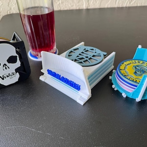 Puede incluir: Tres posavasos impresos en 3D que representan una calavera, un edificio con el texto "Venture Industries" y un posavasos azul y amarillo con el texto "Rusty Pat's Boy Adventures".