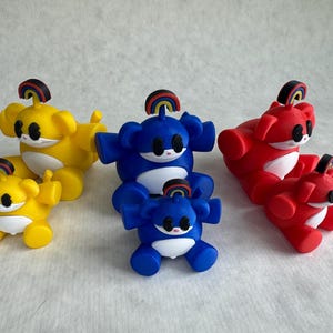 May include: Six small bear-like figurines in yellow, blue, and red, each featuring a white belly, black eyes, and a small rainbow-coloured arch on top of the head. The figures are arranged in pairs of one larger and one smaller size for each colour.