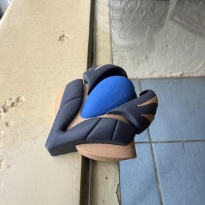 以下が含まれることがあります： 青色のテクスチャード加工された中央部と、黒とタンの幾何学的な形状で構成された3Dプリントオブジェクト。このオブジェクトは、スポーツ用品や機械用の保護コンポーネントのように見えます。