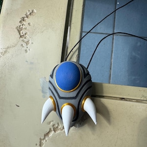 May include: A pendant with a grey body, a blue centre, and three white claw-like protrusions. The pendant has gold accents and black lines. It hangs from a black cord.