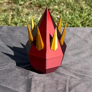May include: A red and gold papercraft crown with a pointed, teardrop shape and six gold spikes. The crown is sitting on a gray surface.