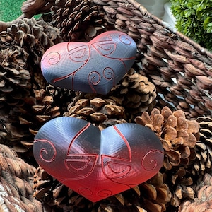 May include: Two heart-shaped ornaments with a red to black gradient. Each heart has an intricate design of curved lines and swirls. The hearts are displayed among pine cones and a woven basket, creating a rustic aesthetic.