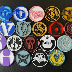 Puede incluir: Una colección de coloridos pines circulares de esmalte con varios diseños. Los pines presentan imágenes como una calavera, una mariposa y texto como "The Venture Bros" y "Rusty's Day Camp For Boy Adventurers".