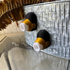 Pode incluir: Dois acessórios decorativos de torneira pretos e amarelos com bicos brancos estão presos a um recipiente de metal galvanizado. Os acessórios têm um design único e são provavelmente para fins decorativos.