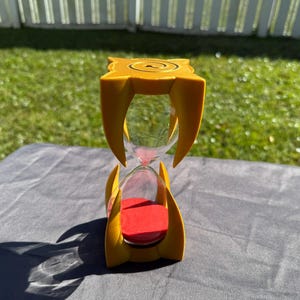 May include: A yellow hourglass with a red sand base and a clear glass bulb. The hourglass is shaped like a claw and has a decorative design on the top.