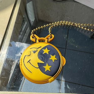 May include: A yellow and blue pendant necklace featuring a smiling crescent moon and three yellow stars. The pendant is round and has a gold-colored chain.