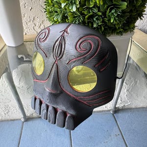 Może przedstawiać: Czarna drewniana maska czaszki z czerwonymi detalami i żółtymi szklanymi oczami. Maska ma misterne rzeźby i zębaty uśmiech.