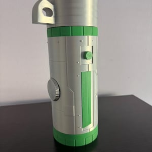 Puede incluir: Un objeto cilíndrico de color plateado con detalles en verde. El objeto tiene un asa, un botón circular y un panel verde. La base y la parte superior también son verdes. El objeto está sobre una superficie oscura.